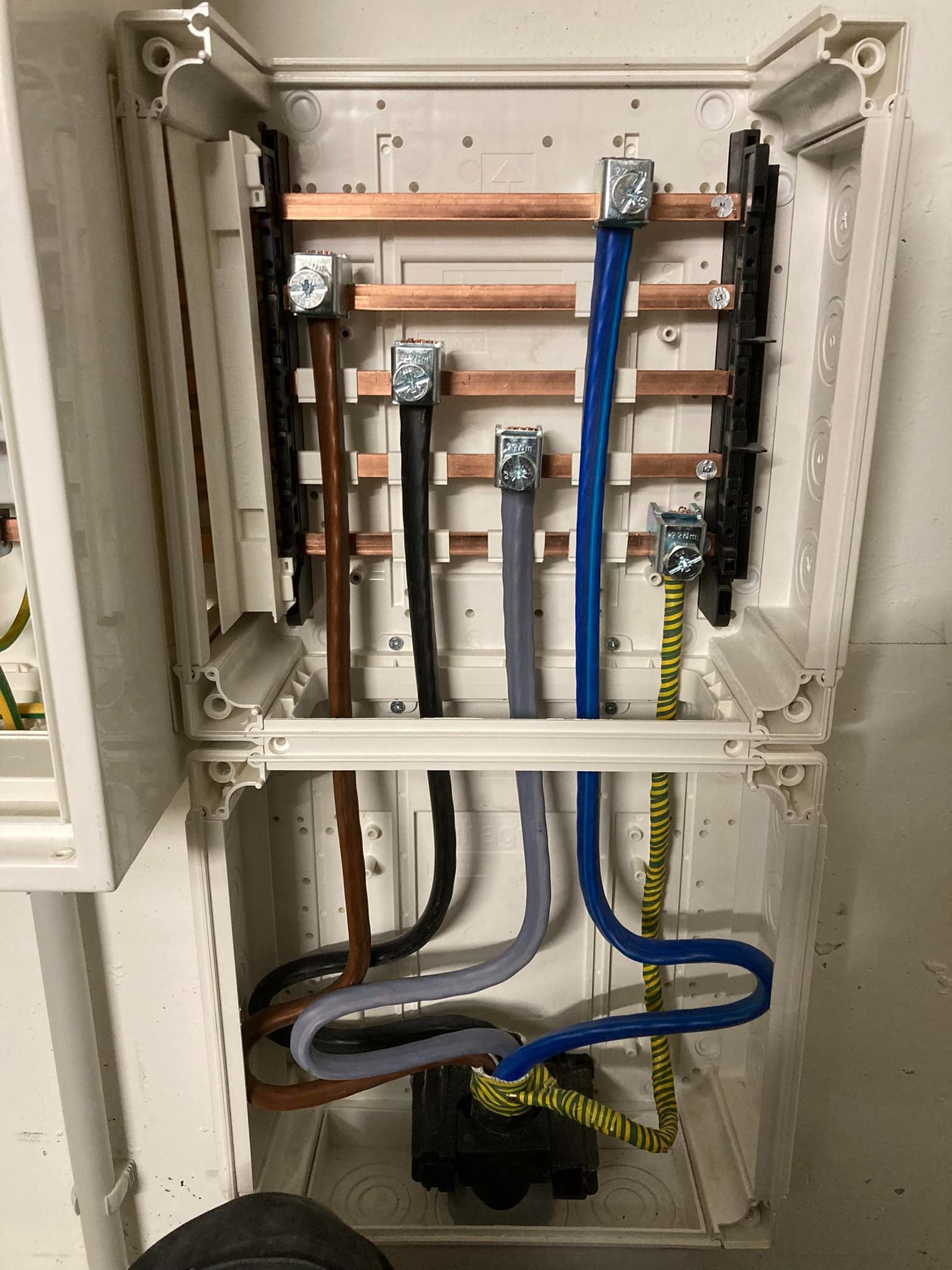 Otwarta skrzynka elektryczna z widocznymi przewodami o różnych kolorach (brązowy, czarny, szary, niebieski, żółto-zielony), podłączonymi do szyn miedzianych i listwy zaciskowej; widoczne ślady...