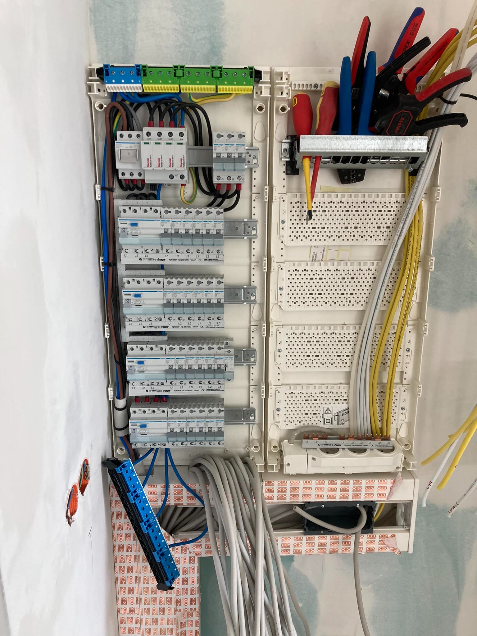 Rozdzielnica elektryczna w trakcie montażu, widoczne zabezpieczenia nadprądowe, różnicowoprądowe, listwy zaciskowe, przewody w kolorach, narzędzia (kombinerki, śrubokręty) umieszczone na specjalnym...
