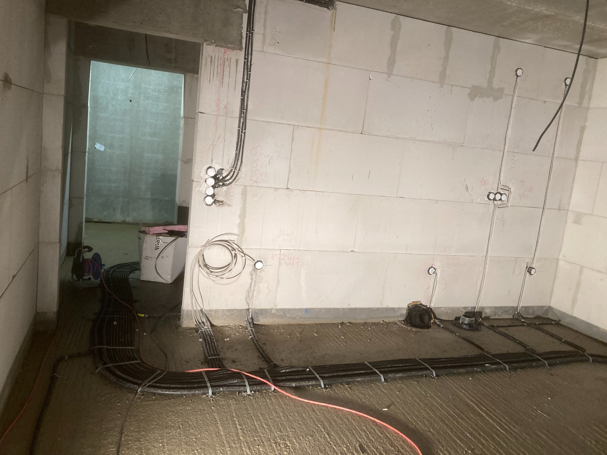 Surowy stan instalacji elektrycznej w nowym budynku: kable w rurach karbowanych ułożone na podłodze, wyprowadzenia przewodów ze ścian z widocznymi puszkami instalacyjnymi, notatki na ścianach.