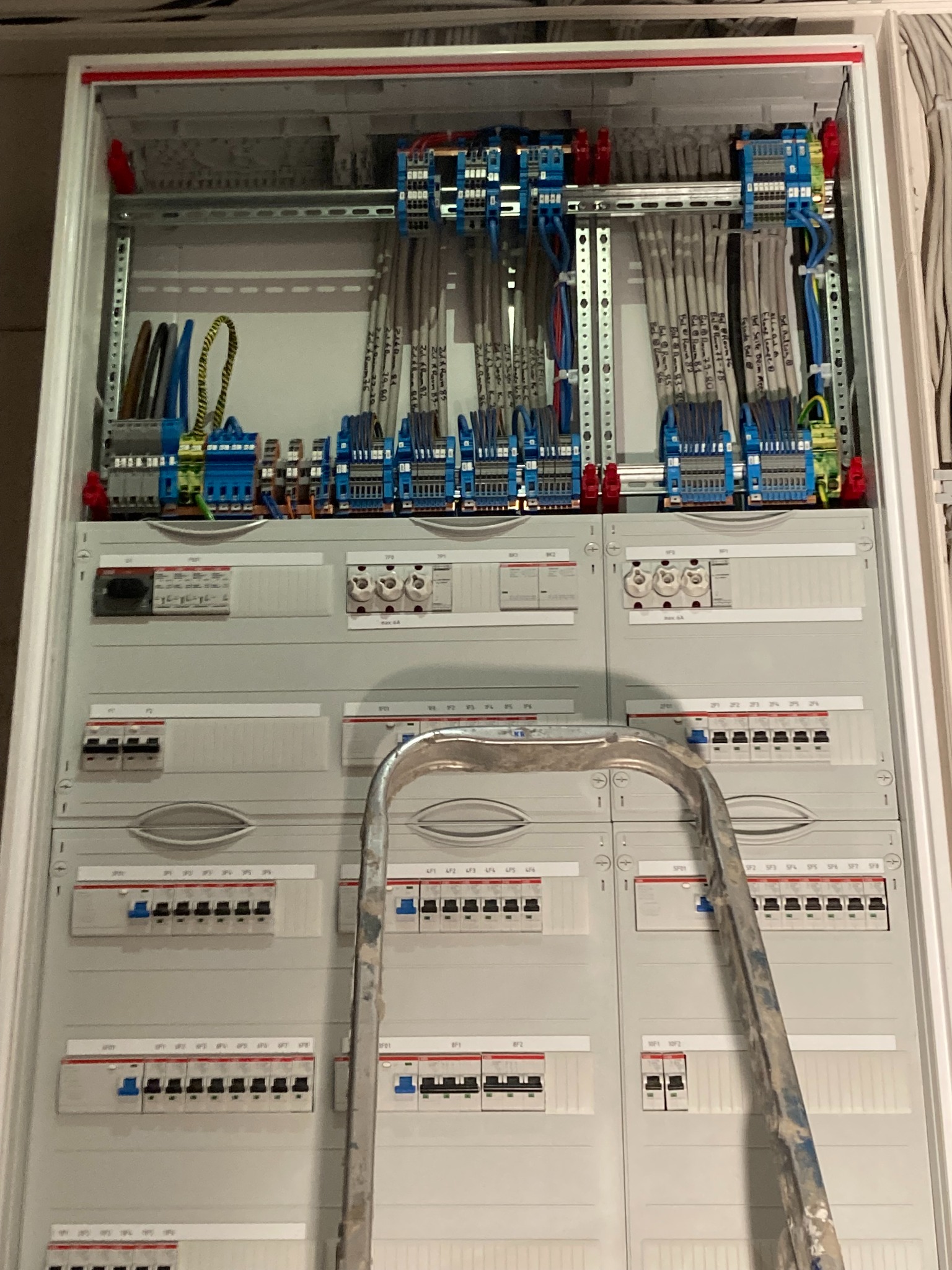 Otwarta szafa rozdzielcza z licznymi przewodami, bezpiecznikami i elementami łączeniowymi, widoczna drabina oparta o szafę, górne partie szafy z napisami na przewodach.