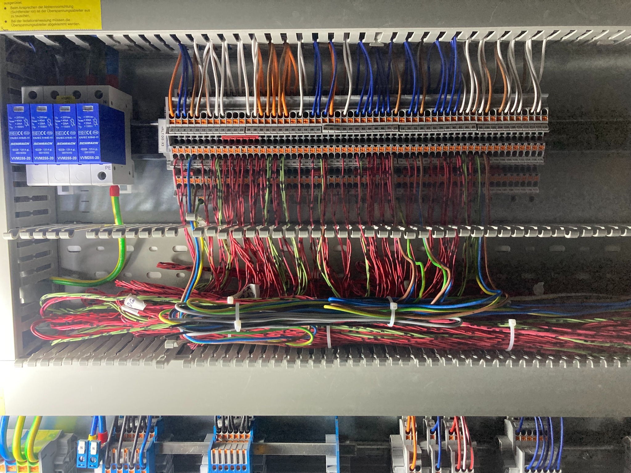 Panel elektryczny z licznymi przewodami o różnych kolorach podłączonymi do listew zaciskowych i zabezpieczeniami przepięciowymi, widoczne etykiety i oznaczenia na elementach.