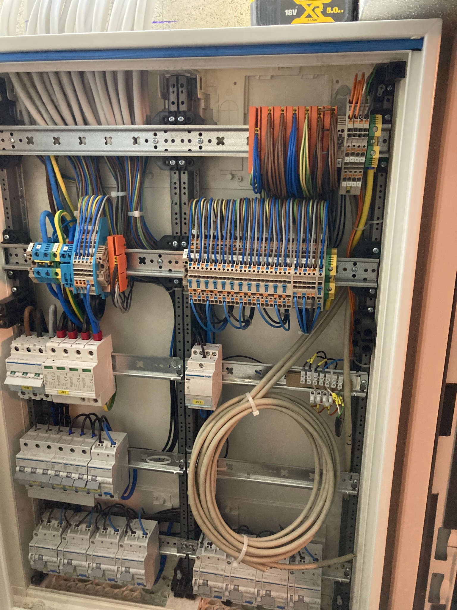 Otwarta szafa elektryczna z widocznymi zabezpieczeniami, przewodami i listwami zaciskowymi, uporządkowane okablowanie, zasilana bateria 18V 5.0Ah na górze.