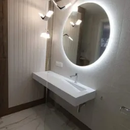 Nowoczesna łazienka z podświetlanym okrągłym lustrem, białą, minimalistyczną umywalką, designerskimi lampami wiszącymi i marmurową podłogą.