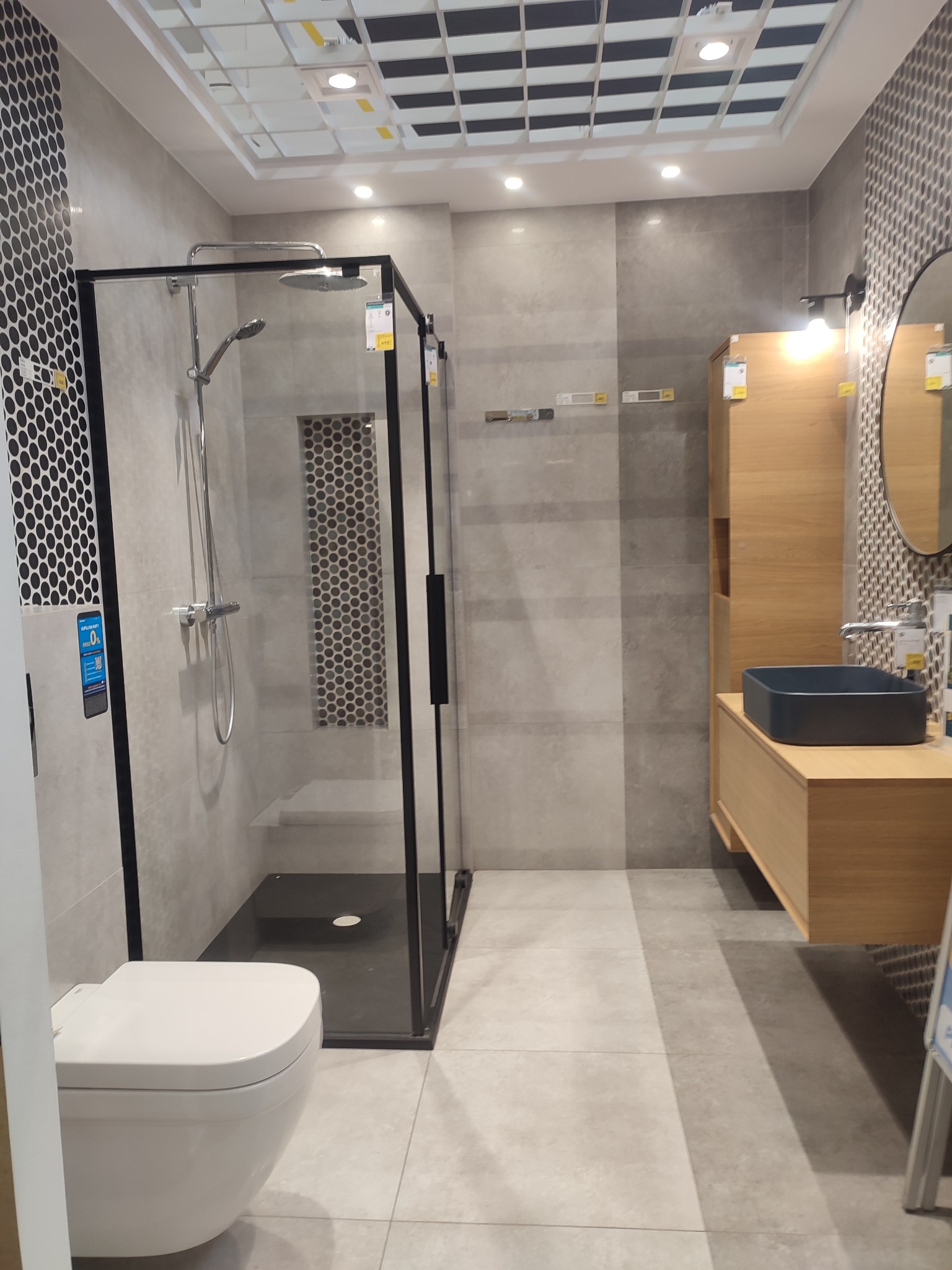 Nowoczesna aranżacja łazienki z kabiną prysznicową typu walk-in z czarnymi profilami, wiszącą toaletą, umywalką nablatową w kolorze grafitowym i szafką z jasnego drewna.