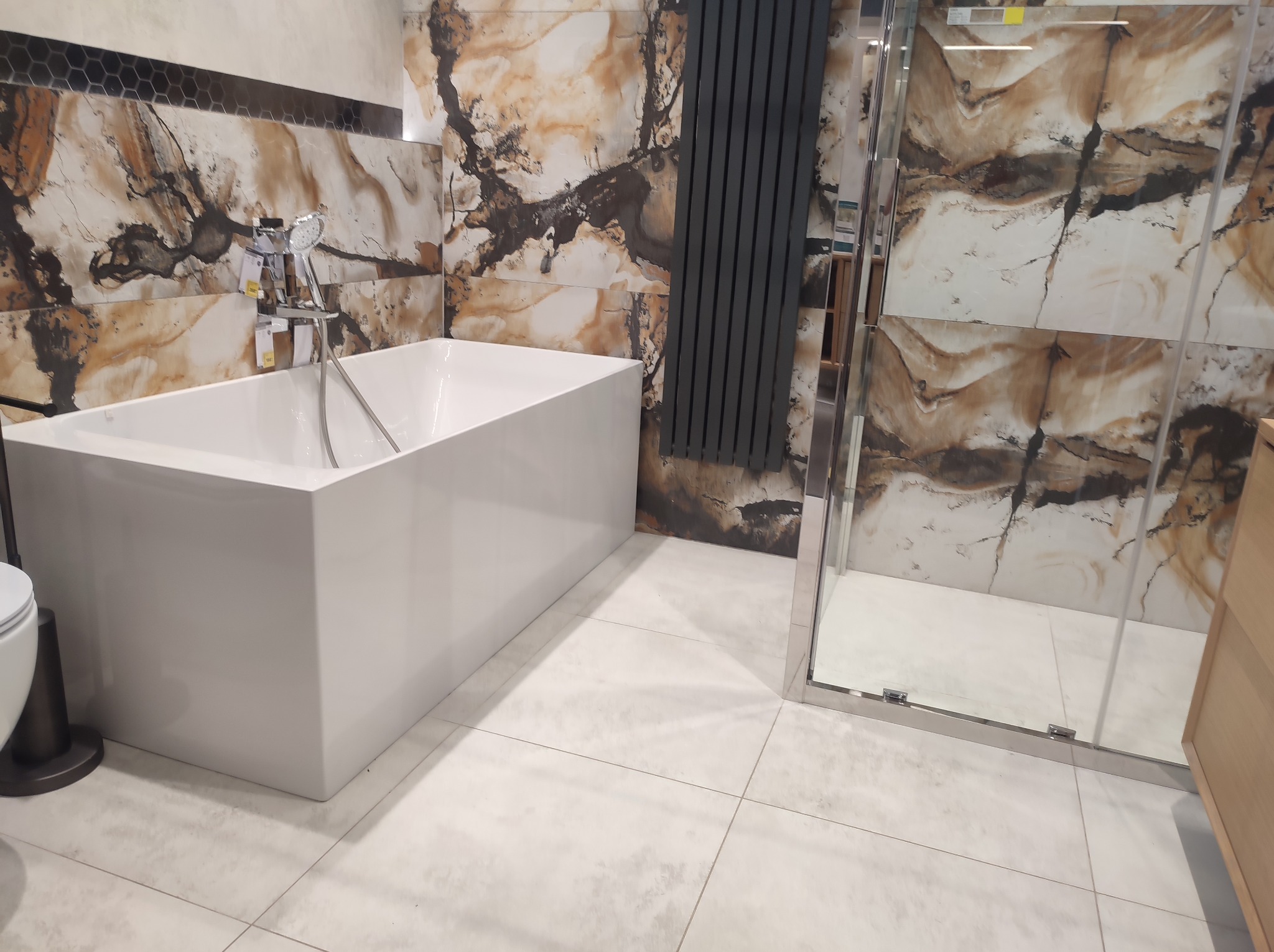 Nowoczesna łazienka z białą, prostokątną wanną, baterią wannową, grzejnikiem drabinkowym w kolorze antracytowym i kabiną prysznicową ze szklanymi drzwiami, wykończona płytkami imitującymi marmur...