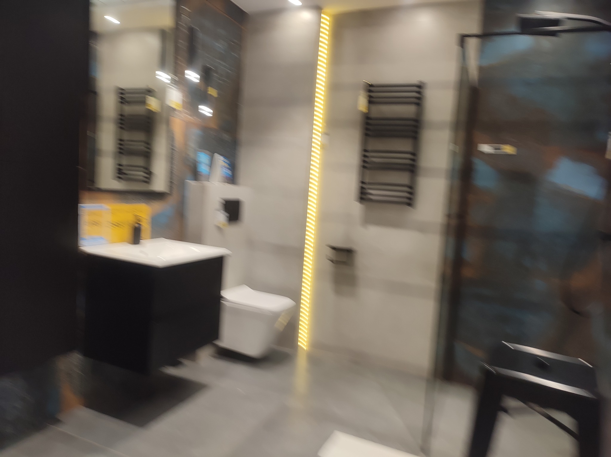 Nowoczesna łazienka z umywalką na czarnej szafce, podwieszaną toaletą, grzejnikiem drabinkowym i kabiną prysznicową z ciemnymi, wzorzystymi panelami.