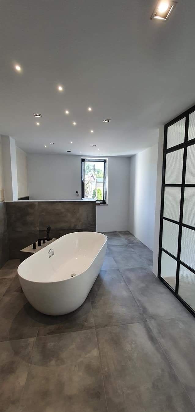 Nowoczesna łazienka z wolnostojącą białą wanną, szarą podłogą z płytek imitujących beton, czarną armaturą i przeszkloną ścianką działową z czarnymi szprosami.