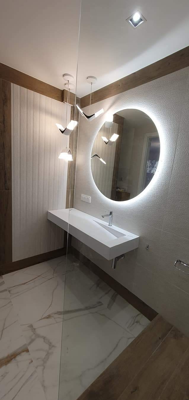 Nowoczesna łazienka z podświetlanym okrągłym lustrem, białą, minimalistyczną umywalką, designerskimi lampami wiszącymi i marmurową podłogą.