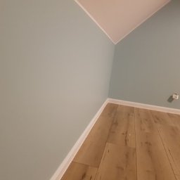 Mal-masters - Pomalowane na pastelowy błękit ściany w pokoju na poddaszu z ułożonymi panelami podłogowymi w odcieniach jasnego drewna, widoczny biały cokół i gniazdko elektryczne.