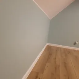 Pomalowane na pastelowy błękit ściany w pokoju na poddaszu z ułożonymi panelami podłogowymi w odcieniach jasnego drewna, widoczny biały cokół i gniazdko elektryczne.