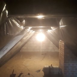 Mal-masters - Poddasze w trakcie prac ociepleniowych, widoczna folia ochronna, ceglany komin i prowizoryczne oświetlenie żarówką.