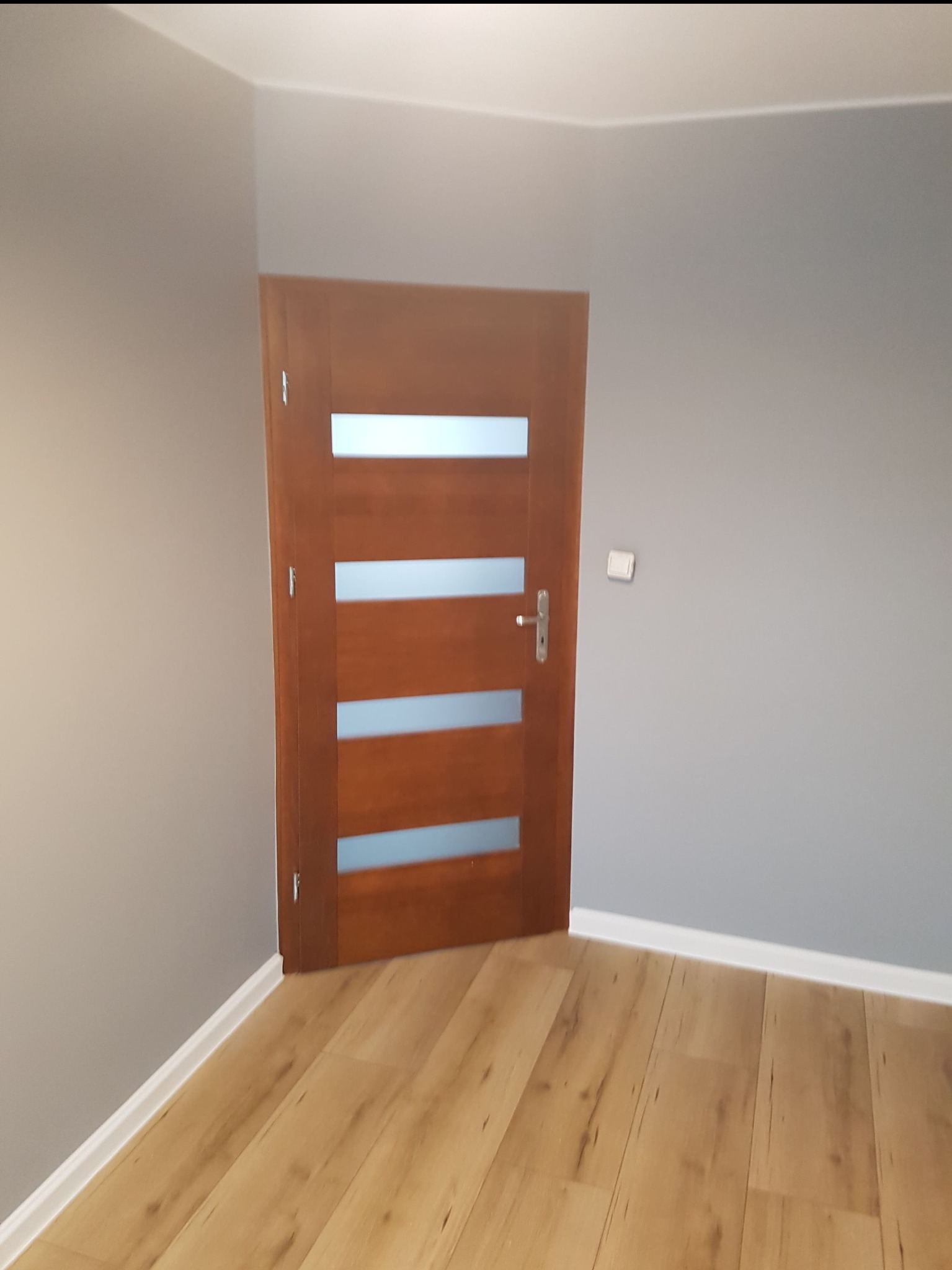 Beżowe panele podłogowe ułożone w pokoju z szarą ścianą i brązowymi drzwiami z czterema poziomymi, mlecznymi szybami.