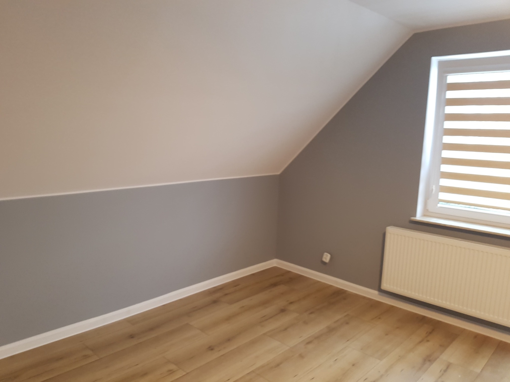 Pomalowane dwukolorowo poddasze ze skosami, z panelami podłogowymi, białym grzejnikiem pod oknem z roletami poziomymi.