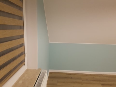 Poddasze po remoncie: ściany pomalowane na dwa kolory - górna część biała, dolna w odcieniach błękitu, drewniane panele podłogowe, roleta okienna w odcieniach brązu.