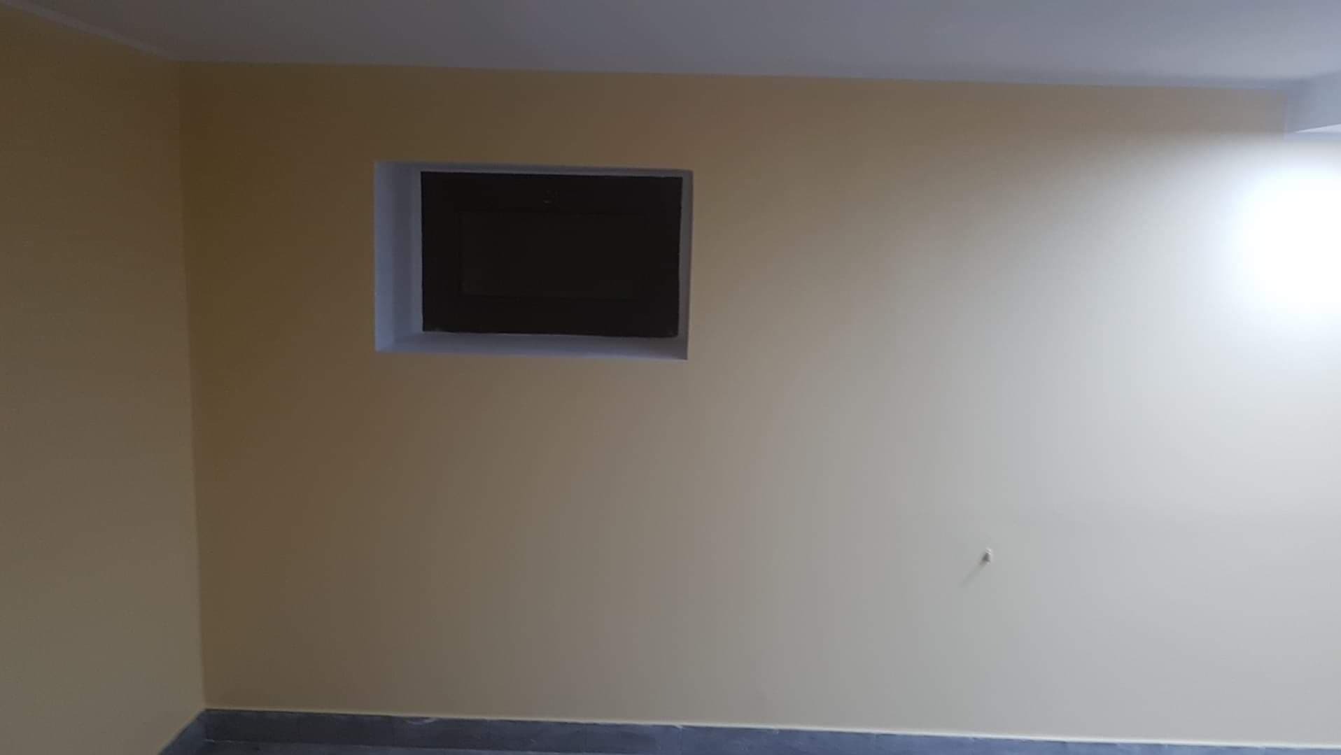 Świeżo pomalowane ściany w pokoju na jasny odcień żółci z ciemnym otworem rewizyjnym, widoczny fragment szarej podłogi.