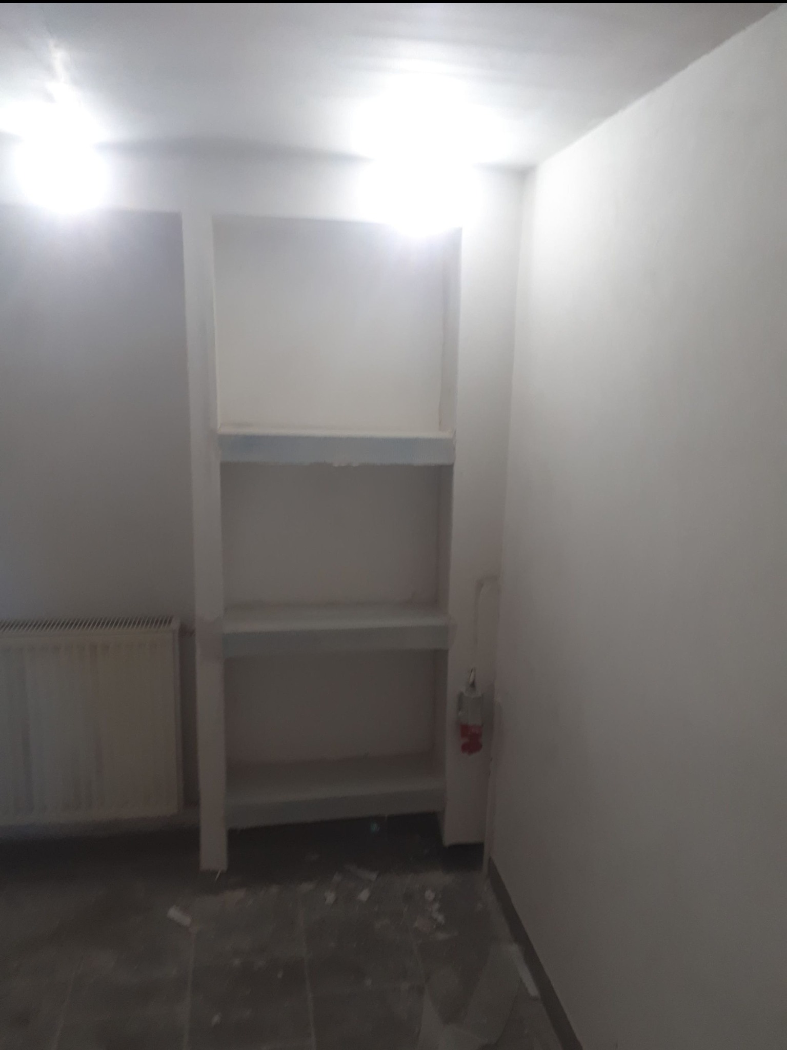 Wnętrze w trakcie remontu: białe ściany, wnęka z półkami w surowym stanie, widoczne ślady po pracach na podłodze, grzejnik i instalacja elektryczna.