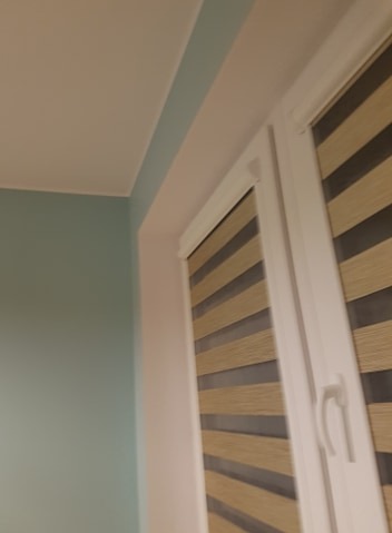 Turkusowa ściana pomalowana w rogu pokoju, obok okno z beżowo-szarymi roletami w białem ramie.