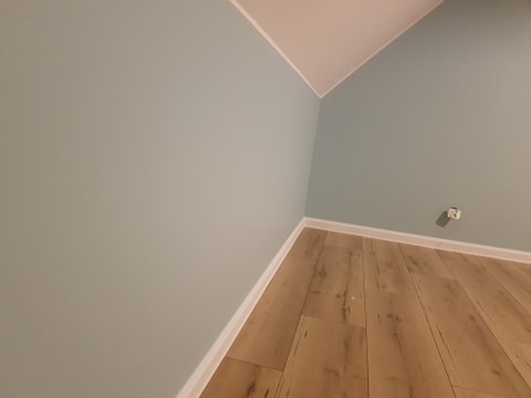 Pomalowane na pastelowy błękit ściany w pokoju na poddaszu z ułożonymi panelami podłogowymi w odcieniach jasnego drewna, widoczny biały cokół i gniazdko elektryczne.