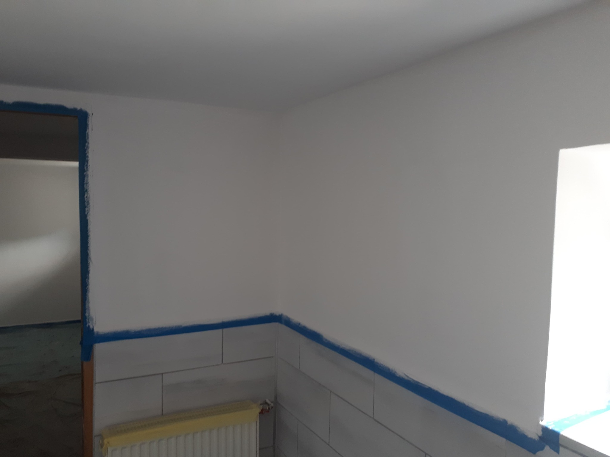 Przygotowane do malowania wnętrze: ściany zabezpieczone niebieską taśmą malarską, widoczne płytki ceramiczne na dole ściany i zakryty grzejnik.