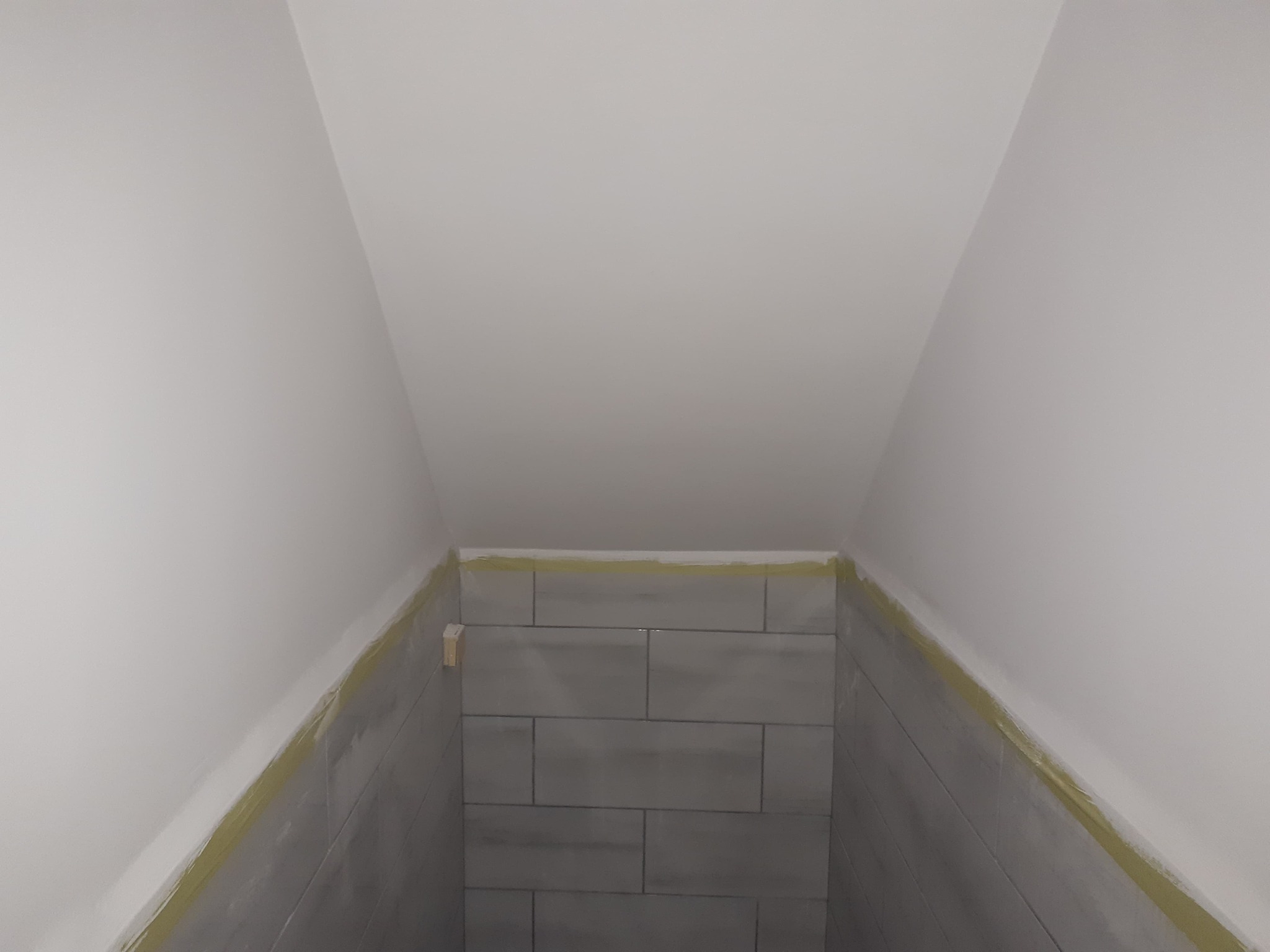 Świeżo położone, szare płytki w wąskim pomieszczeniu pod skosem, zabezpieczone taśmą malarską przy suficie i ścianach, widoczny fragment drewnianego elementu konstrukcyjnego.