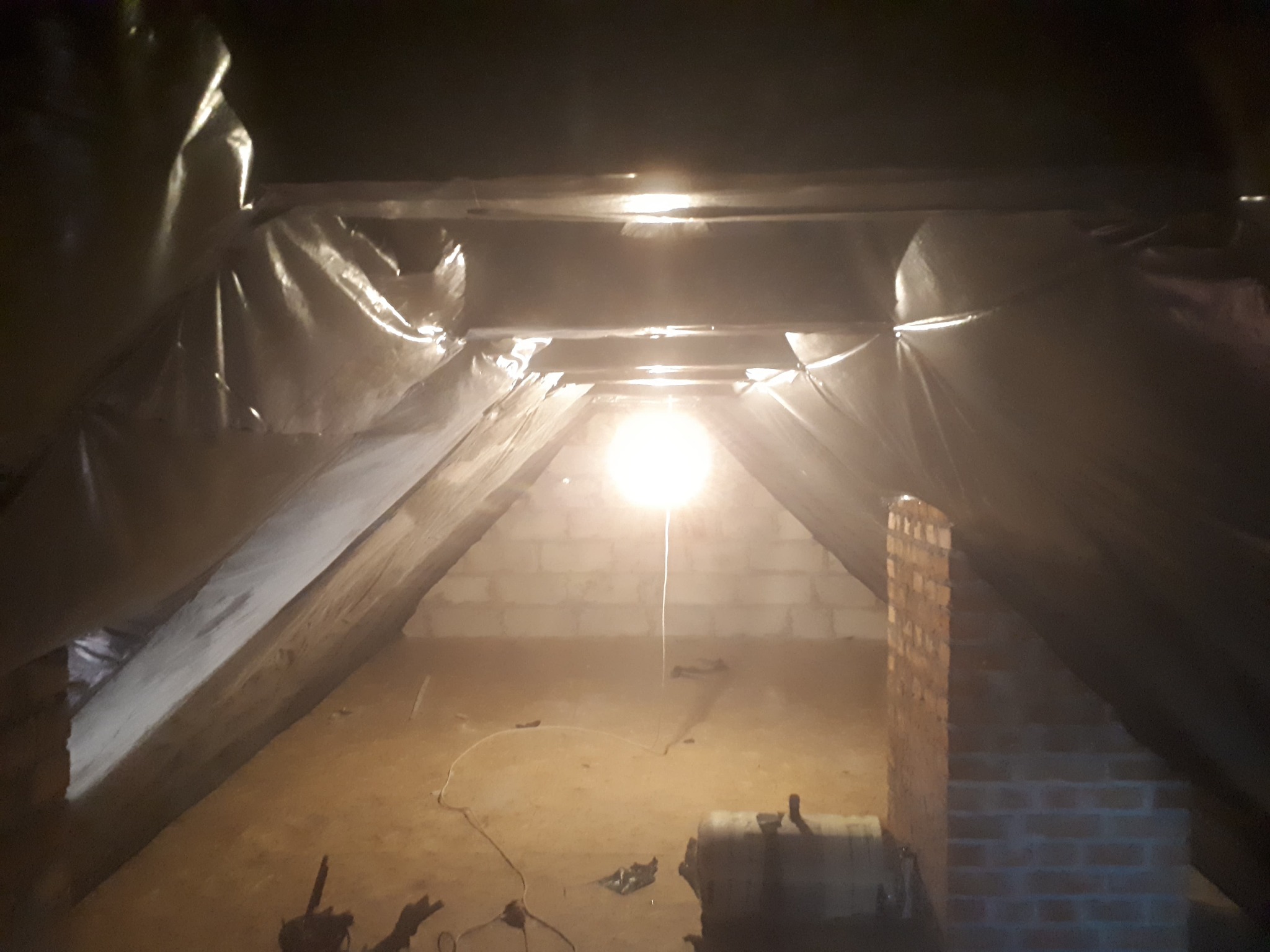 Poddasze w trakcie prac ociepleniowych, widoczna folia ochronna, ceglany komin i prowizoryczne oświetlenie żarówką.