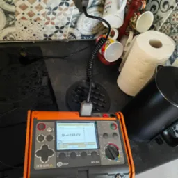 Pomarańczowo-szary miernik elektryczny Sonel MPI-525 z wyświetlaczem wskazującym napięcie 243.7V, ustawiony na czarnym blacie kuchennym obok czajnika i pojemnika na ręczniki papierowe, w tle...