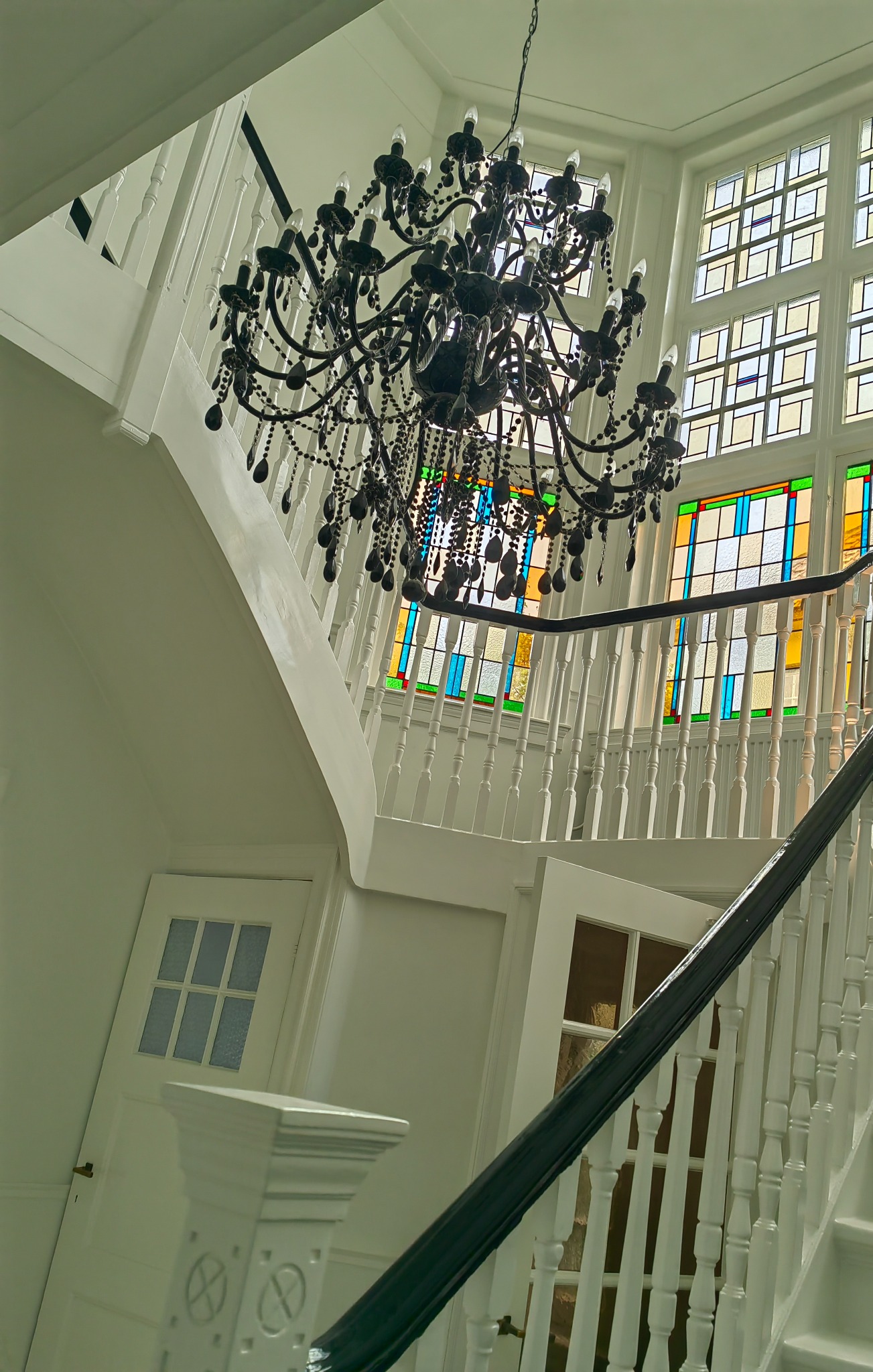 Klatka schodowa z białymi schodami i balustradą, czarną poręczą, oraz ozdobnym czarnym żyrandolem z kryształkami, na tle okien z kolorowymi szybami.