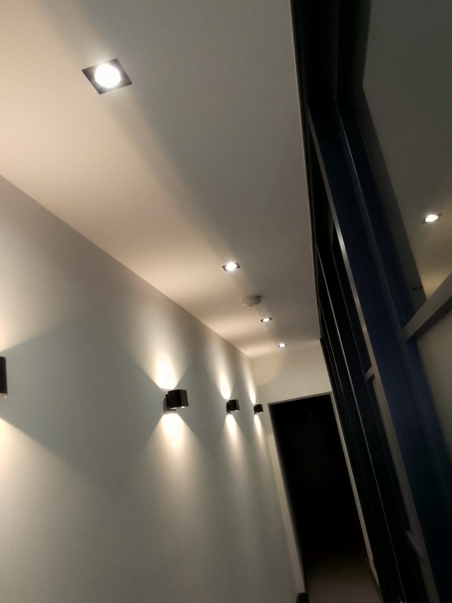 Korytarz oświetlony szeregiem czarnych kinkietów ściennych i wpuszczanych reflektorów sufitowych, widok z dołu, z fragmentem konstrukcji stalowej po prawej.