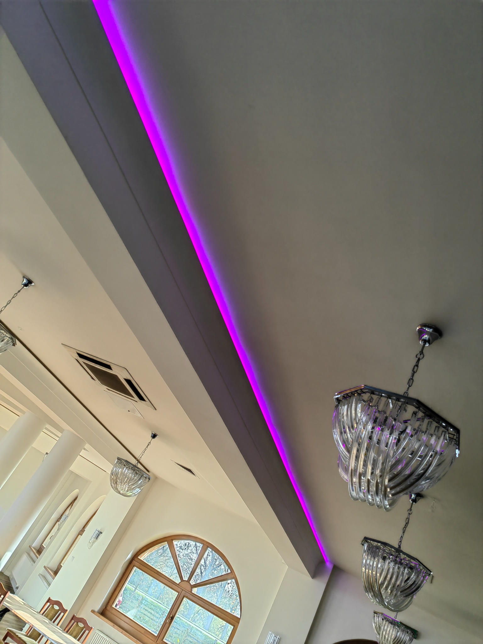 Nowoczesne oświetlenie sufitowe z purpurową listwą LED i kryształowymi żyrandolami w przestronnym wnętrzu z dużymi oknami.