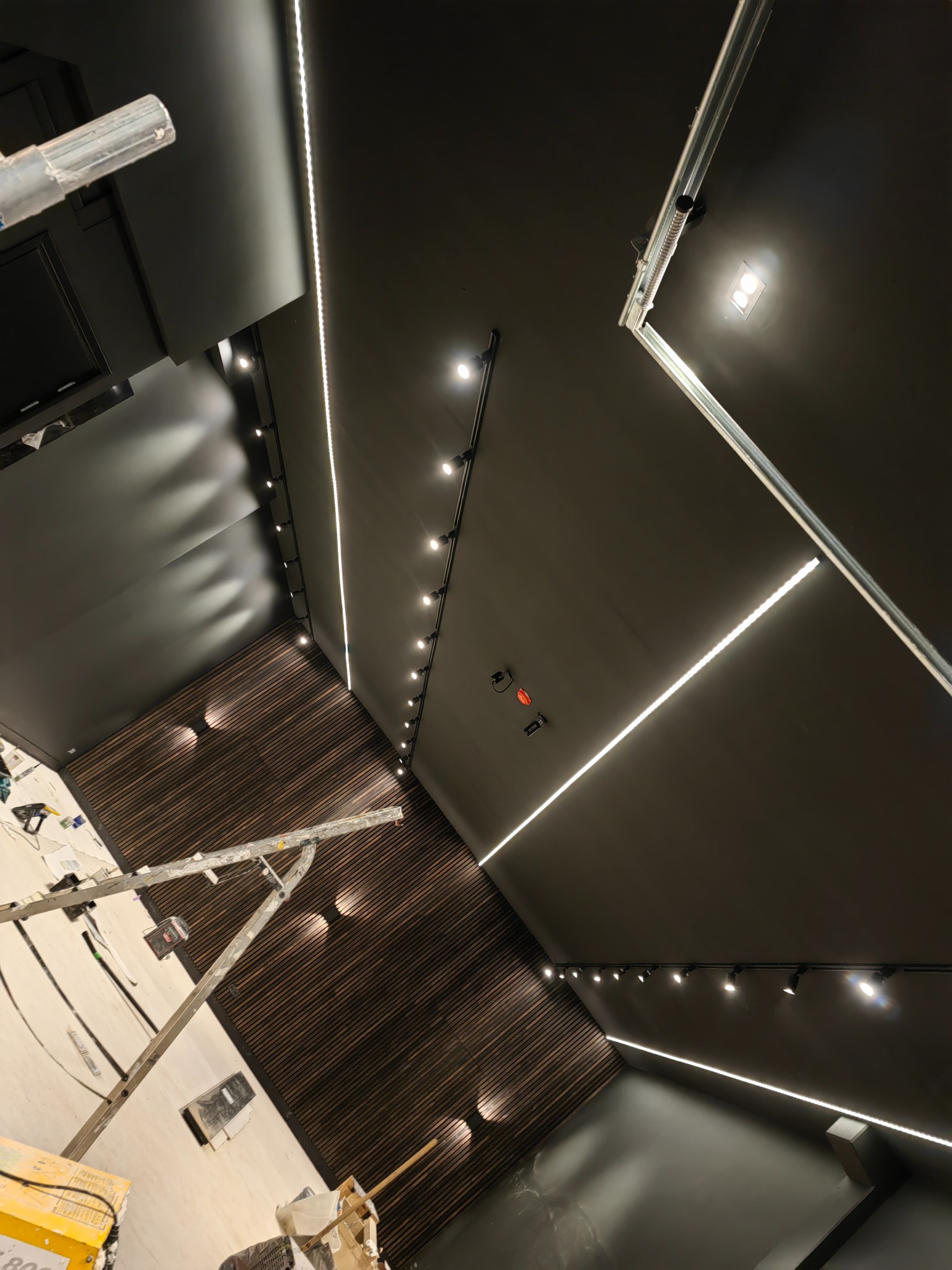 Czarne wnętrze z drewnianą ścianą, oświetlone liniowymi lampami LED i reflektorami na szynoprzewodzie. Widoczny fragment drabiny malarskiej i narzędzia sugerują etap prac wykończeniowych.