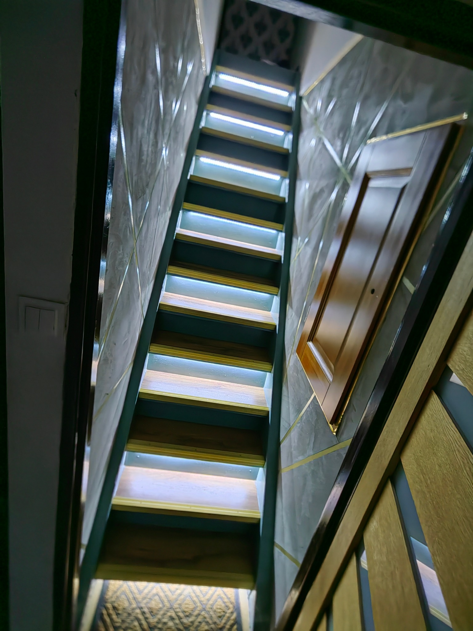 Nowoczesne schody z podświetleniem LED na każdym stopniu, szara ściana z dekoracyjnymi złotymi paskami, widok z dołu.
