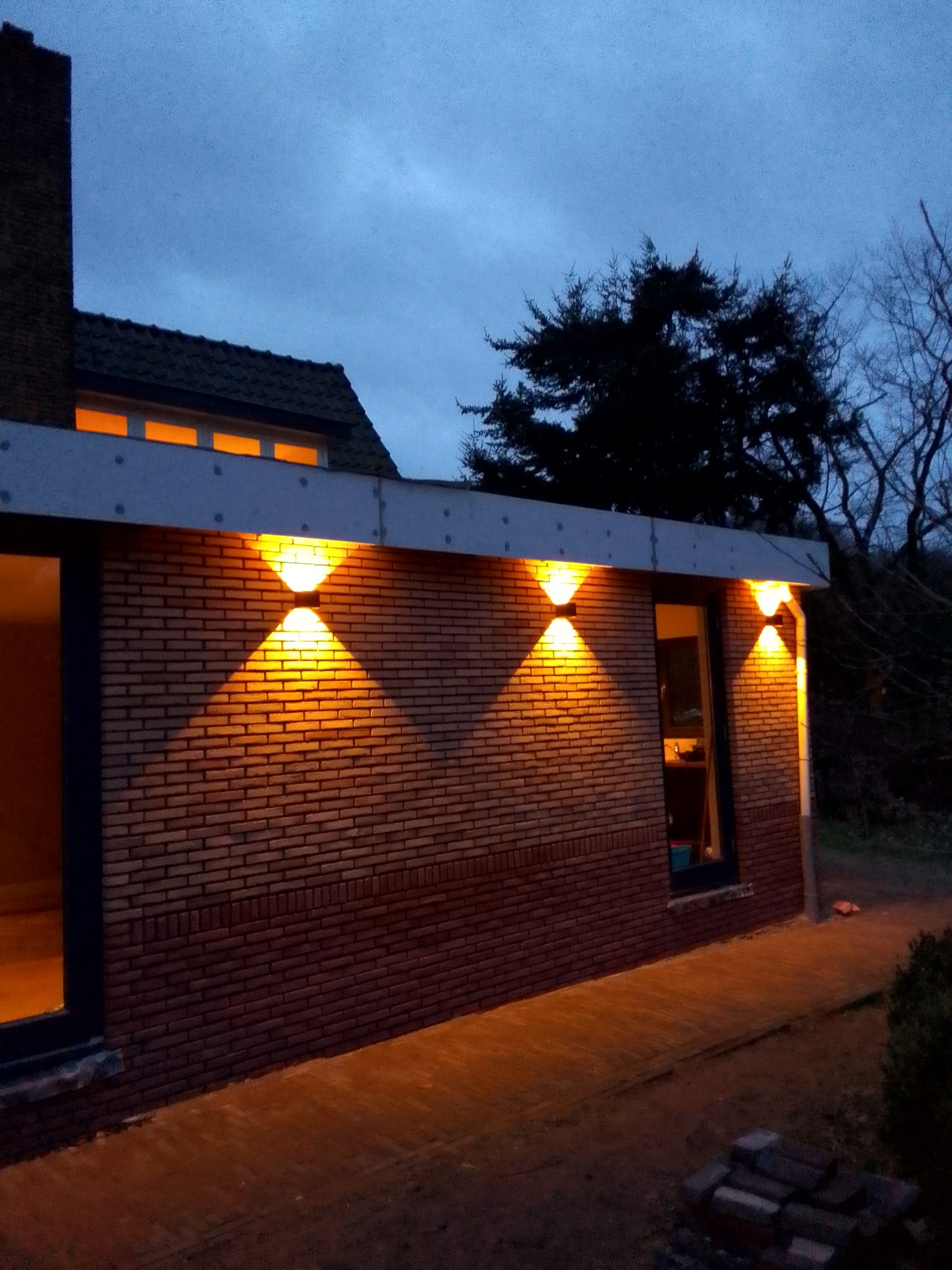 Zewnętrzne oświetlenie budynku z cegły, trzy podwójne lampy ścienne emitujące ciepłe światło w pochmurny wieczór.