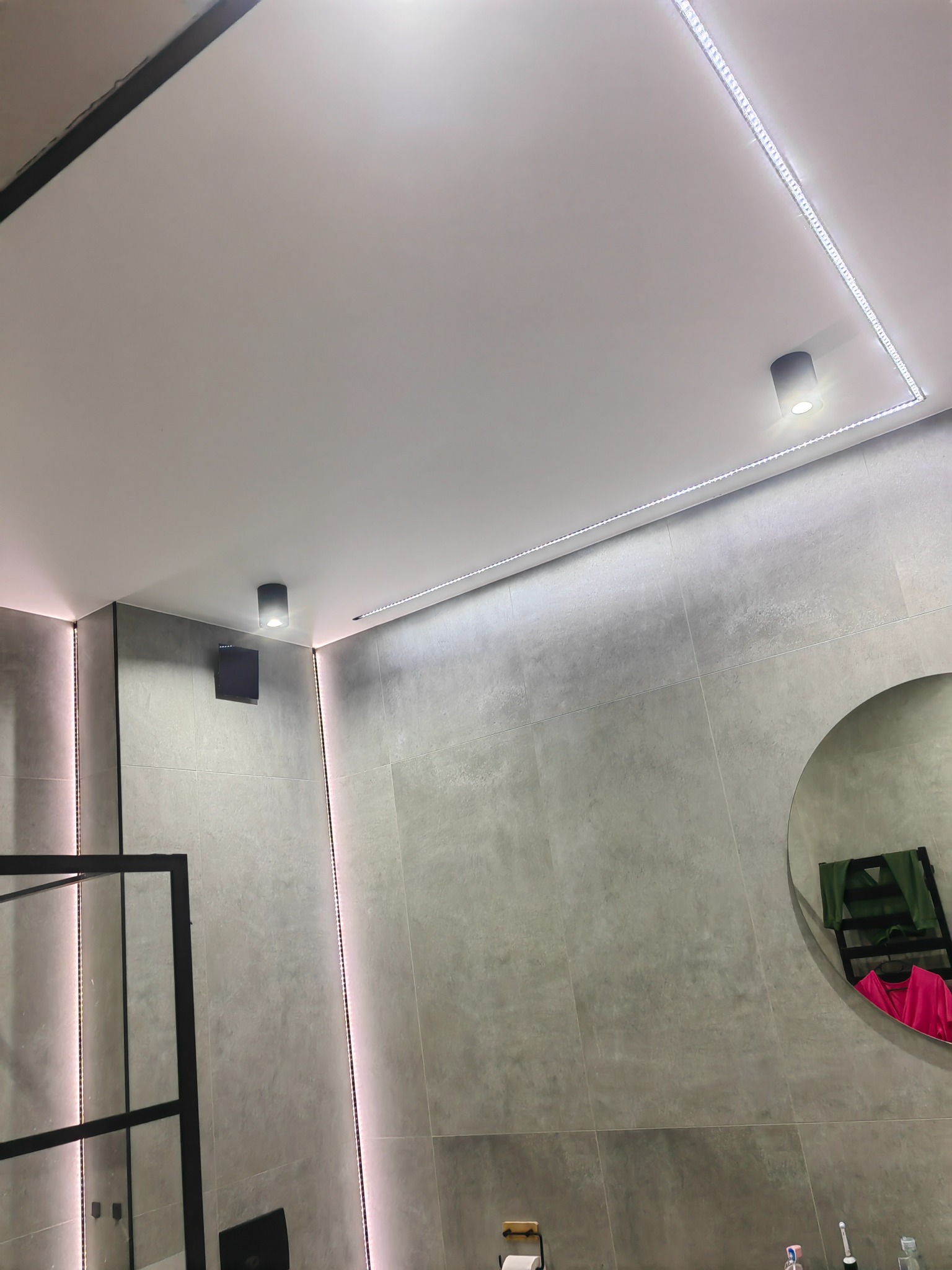 Nowoczesna łazienka z szarymi płytkami, podświetleniem LED na suficie i ścianach oraz okrągłym lustrem.