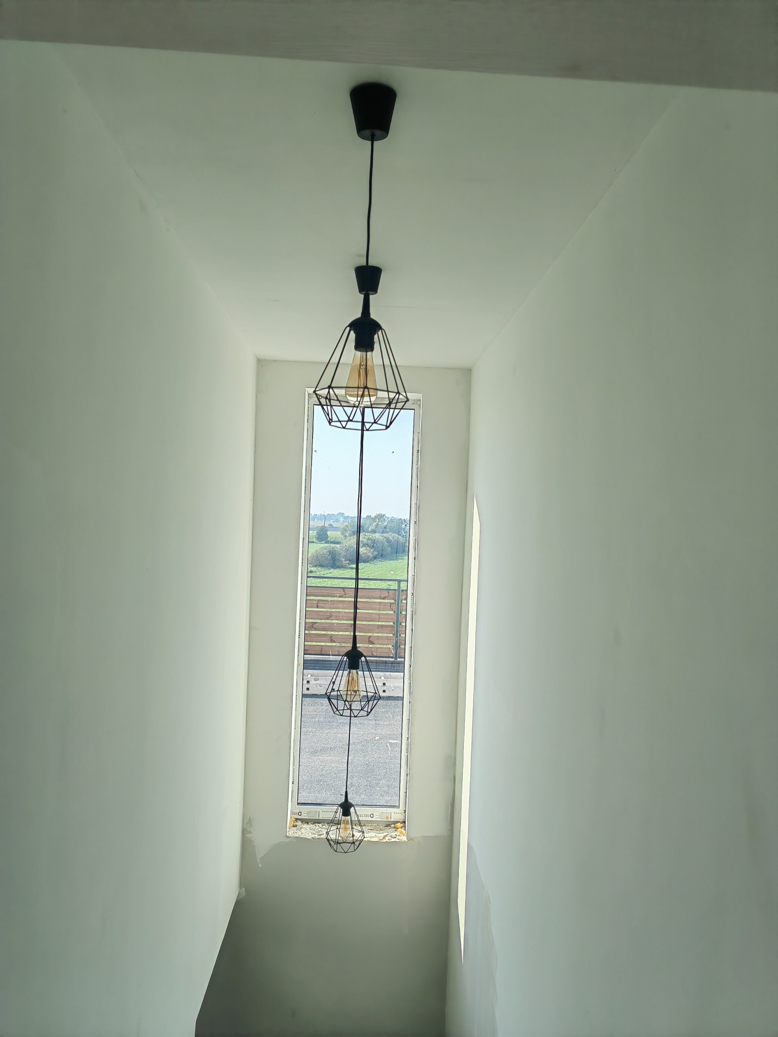 Nowoczesna, czarna lampa wisząca z trzema kloszami w kształcie geometrycznych klatek, zamontowana w jasnym pomieszczeniu z oknem, przez które widać zielony krajobraz.