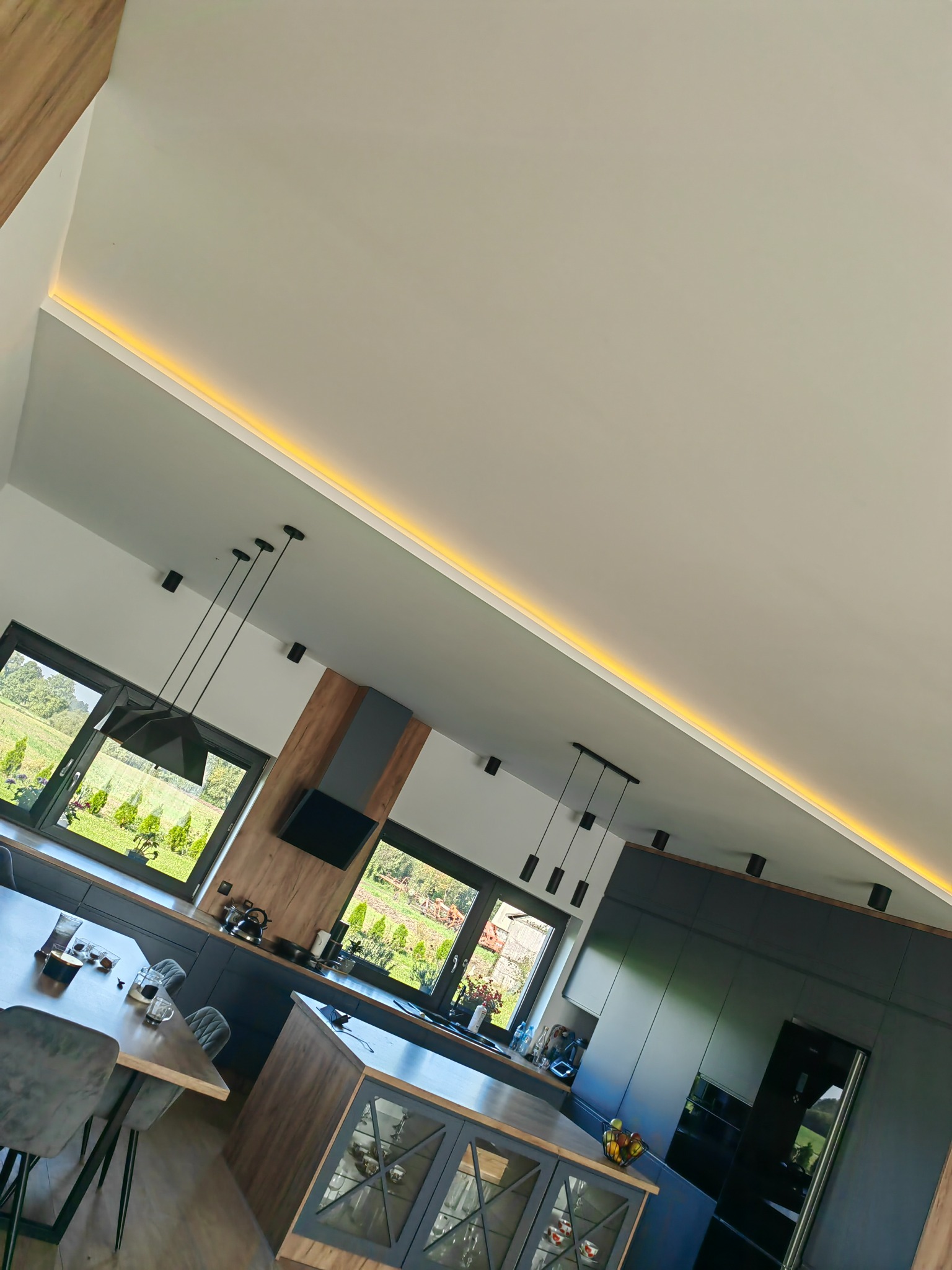 Nowoczesna kuchnia z wyspą, oświetleniem LED podsufitowym i wiszącymi lampami, widok z dołu, okna z widokiem na ogród.