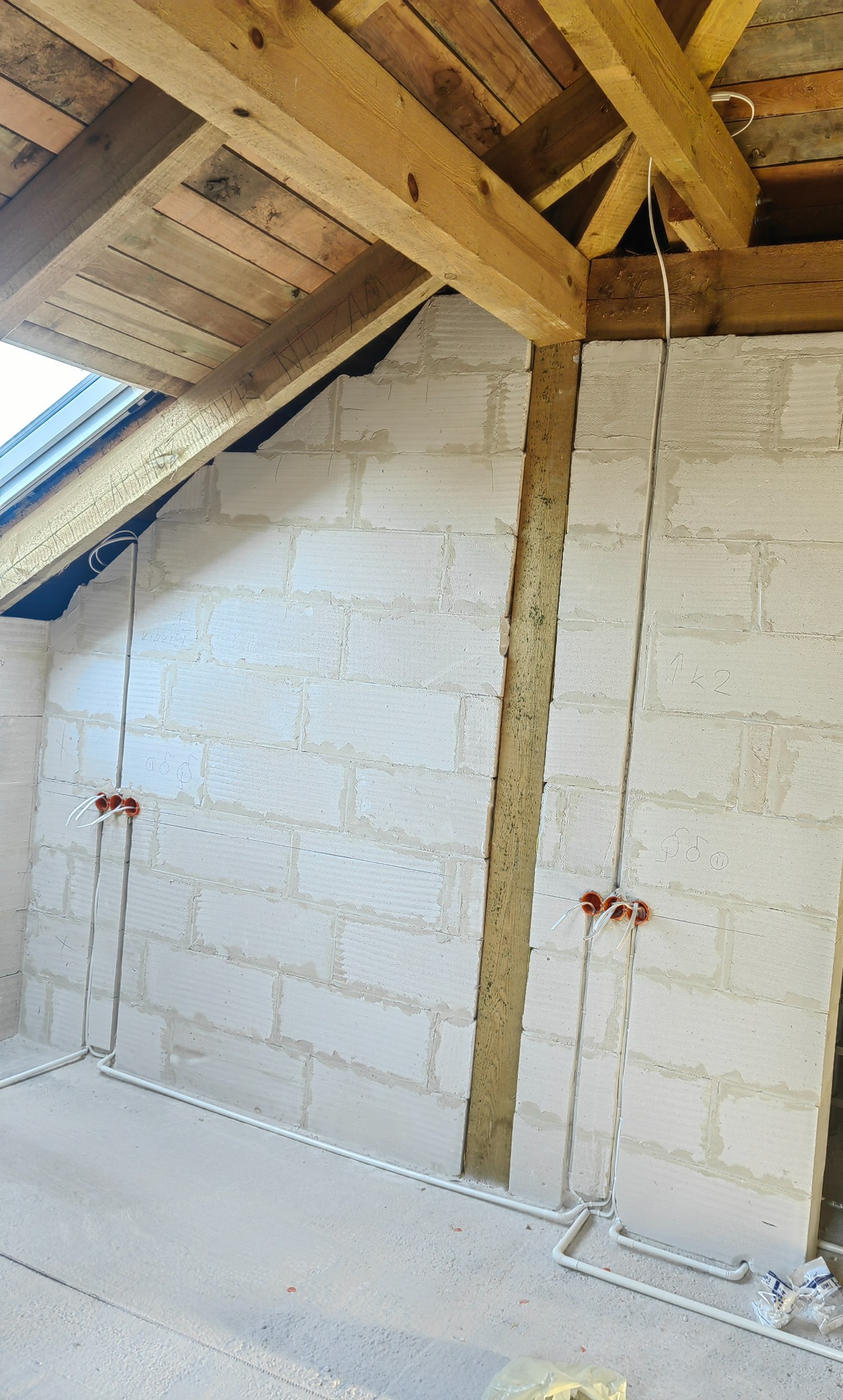 Surowy stan poddasza z widocznymi przewodami elektrycznymi w peszlach, prowadzącymi do puszek instalacyjnych w ścianie z betonu komórkowego, drewniana konstrukcja dachu.