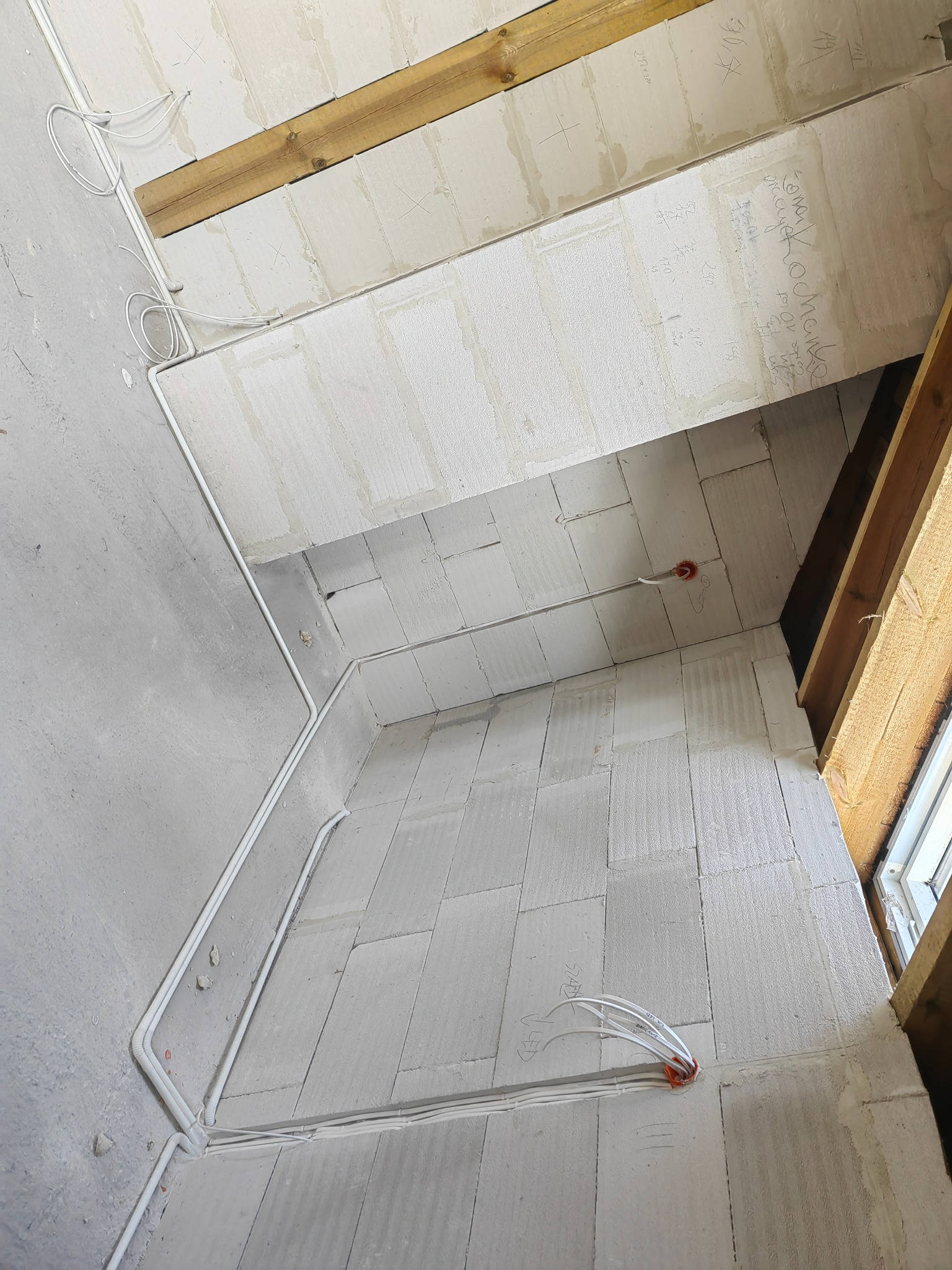 Wnętrze w trakcie prac wykończeniowych; widoczne białe przewody elektryczne poprowadzone po ścianach z białych bloczków, drewniane belki stropowe i okno w rogu, ściany z surowego betonu.