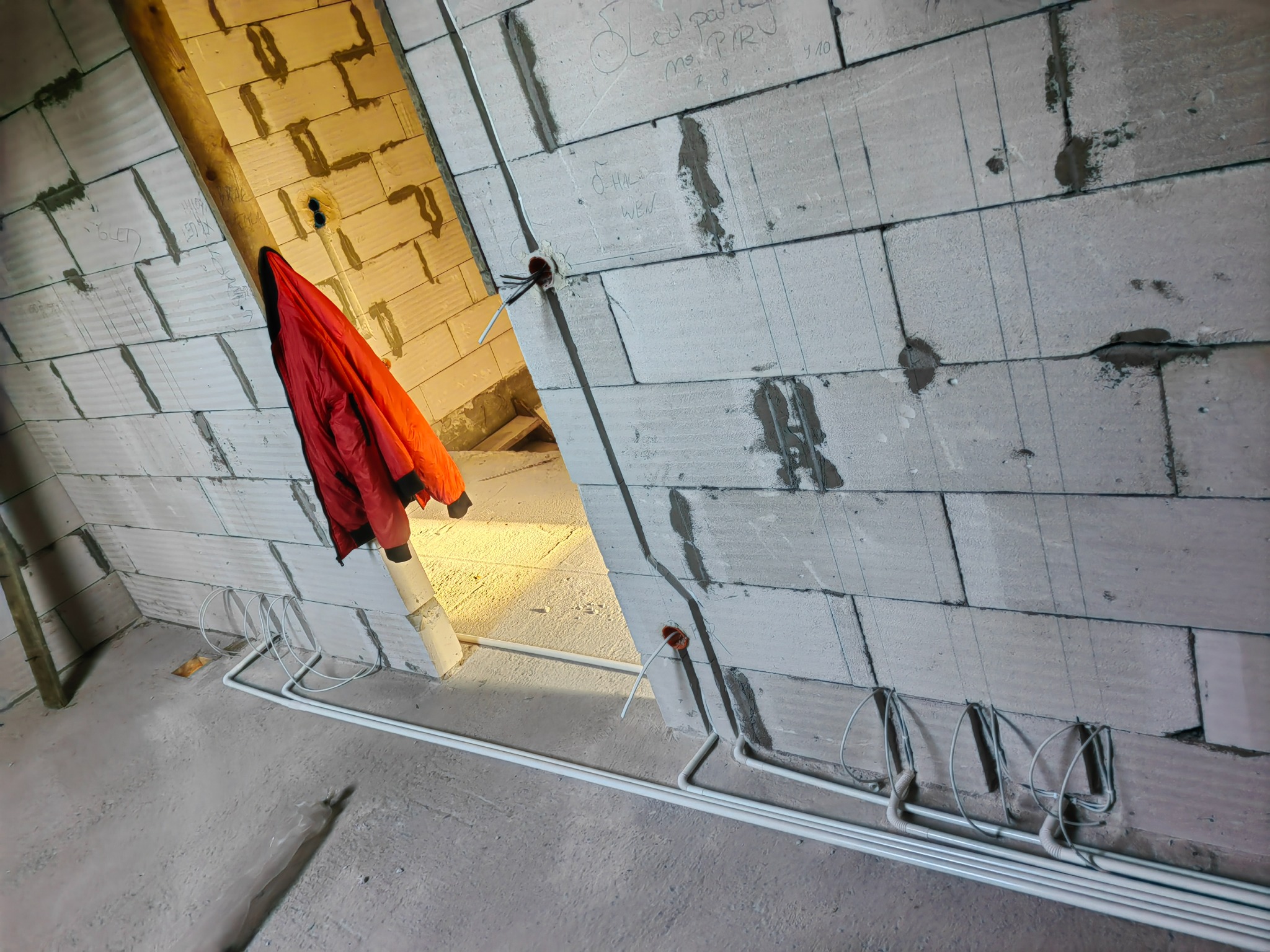 Ściana z betonu komórkowego w trakcie prac, widoczne kable elektryczne w peszlu, otwory pod puszki, oznaczenia na ścianie, czerwona kurtka robocza wisząca w przejściu.