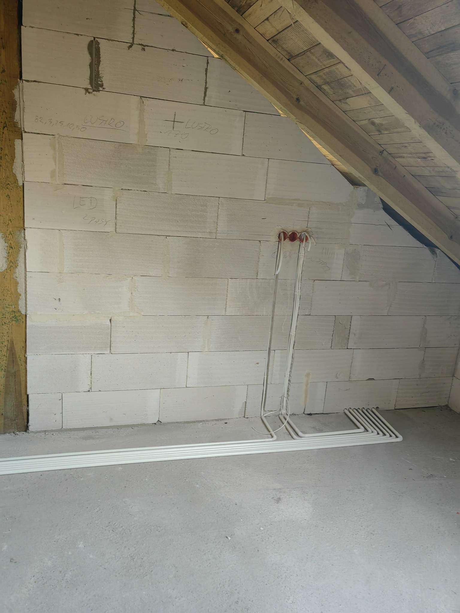 Instalacja elektryczna w surowym stanie poddasza: kable wychodzące z puszki, rury instalacyjne ułożone na podłodze, ściana z bloczków z odręcznymi notatkami o rozmieszczeniu oświetlenia.