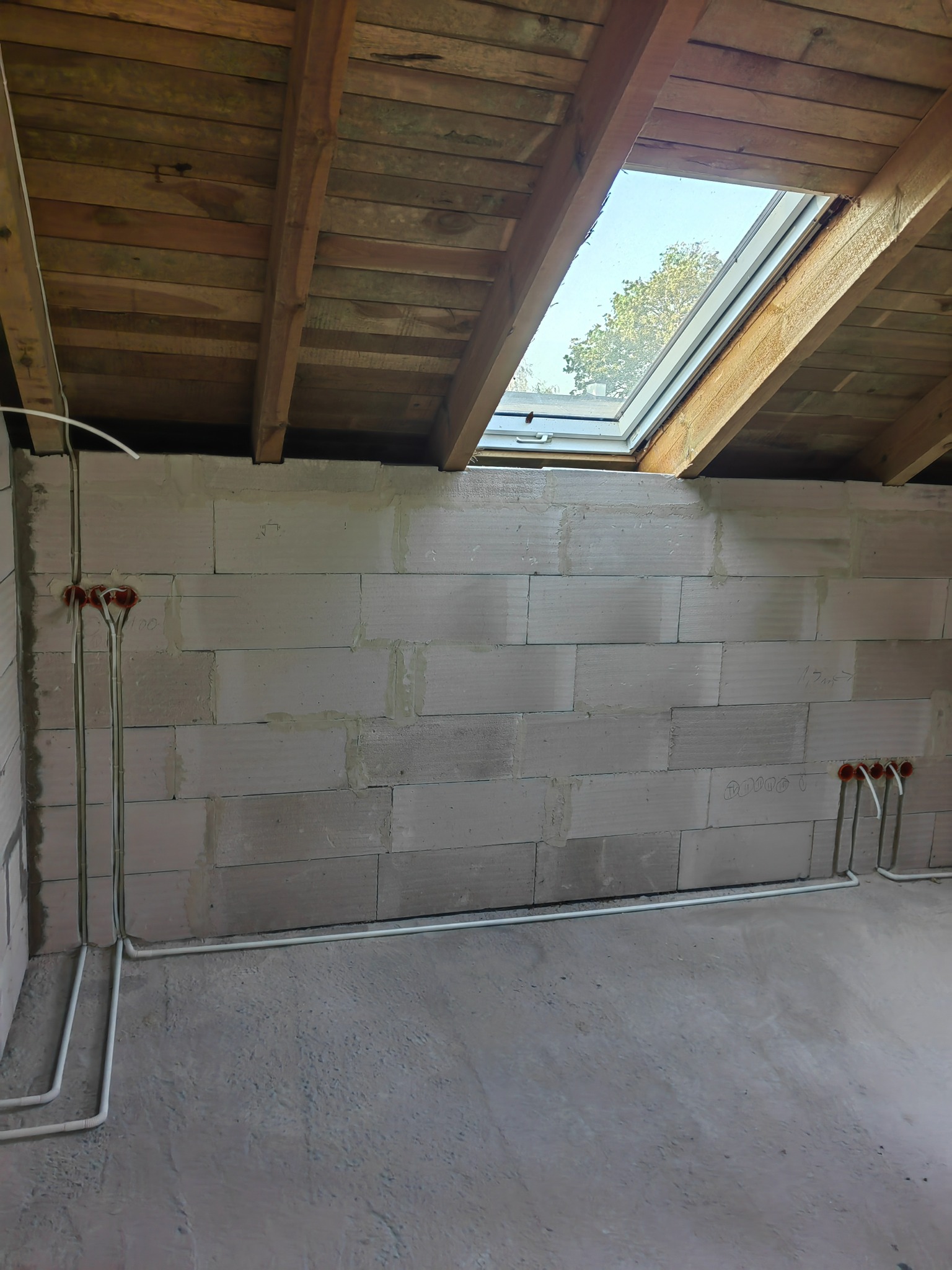 Wewnętrzna instalacja elektryczna w surowym pomieszczeniu z oknem dachowym, widoczne kable w białych rurkach PCV prowadzone po ścianach z betonu komórkowego, puszki elektryczne w kolorze...