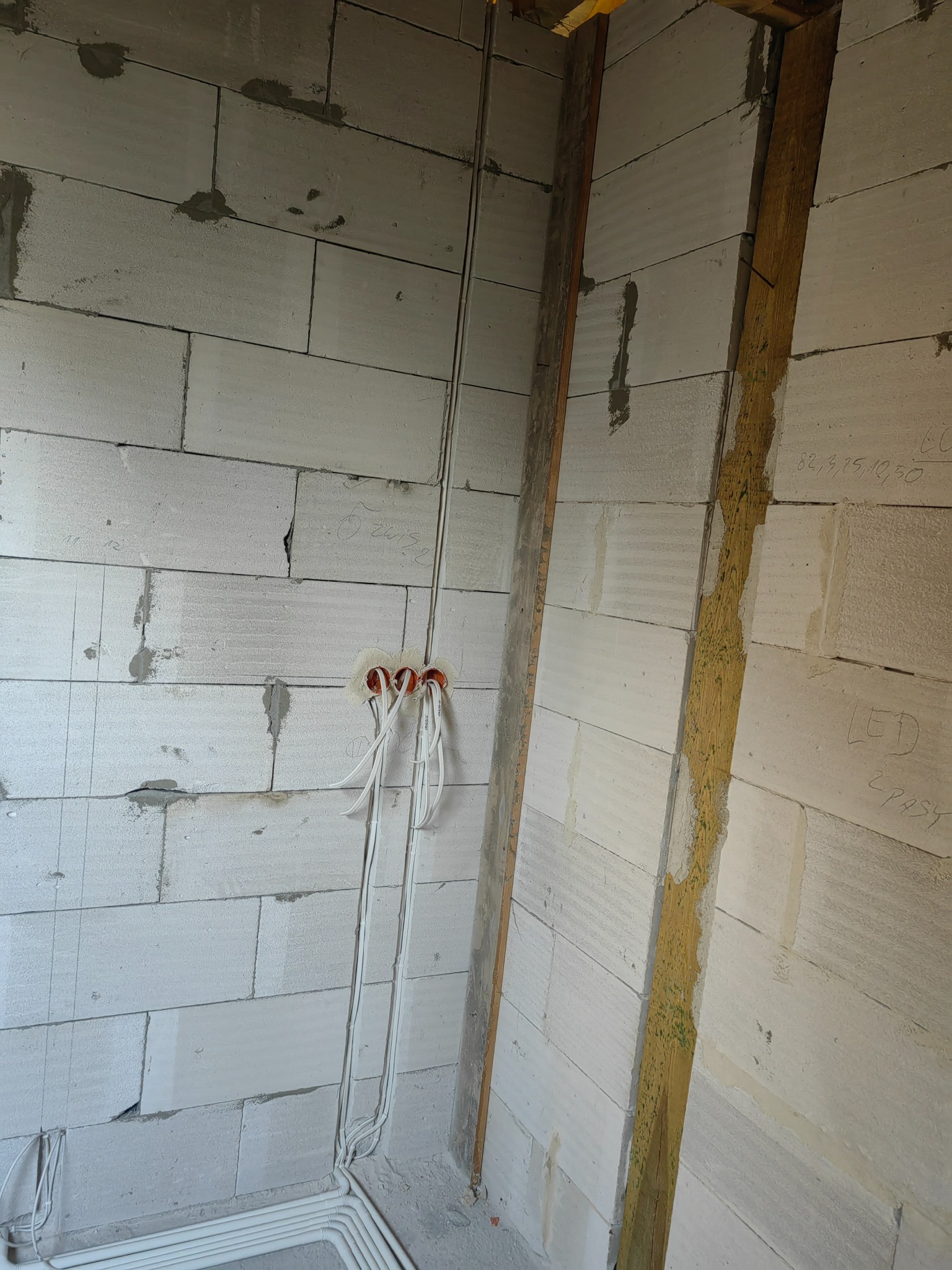 Surowa ściana z betonu komórkowego z trzema puszkami elektrycznymi i wystającymi przewodami, widoczne również rury instalacyjne przy podłodze, w tle drewniana konstrukcja.