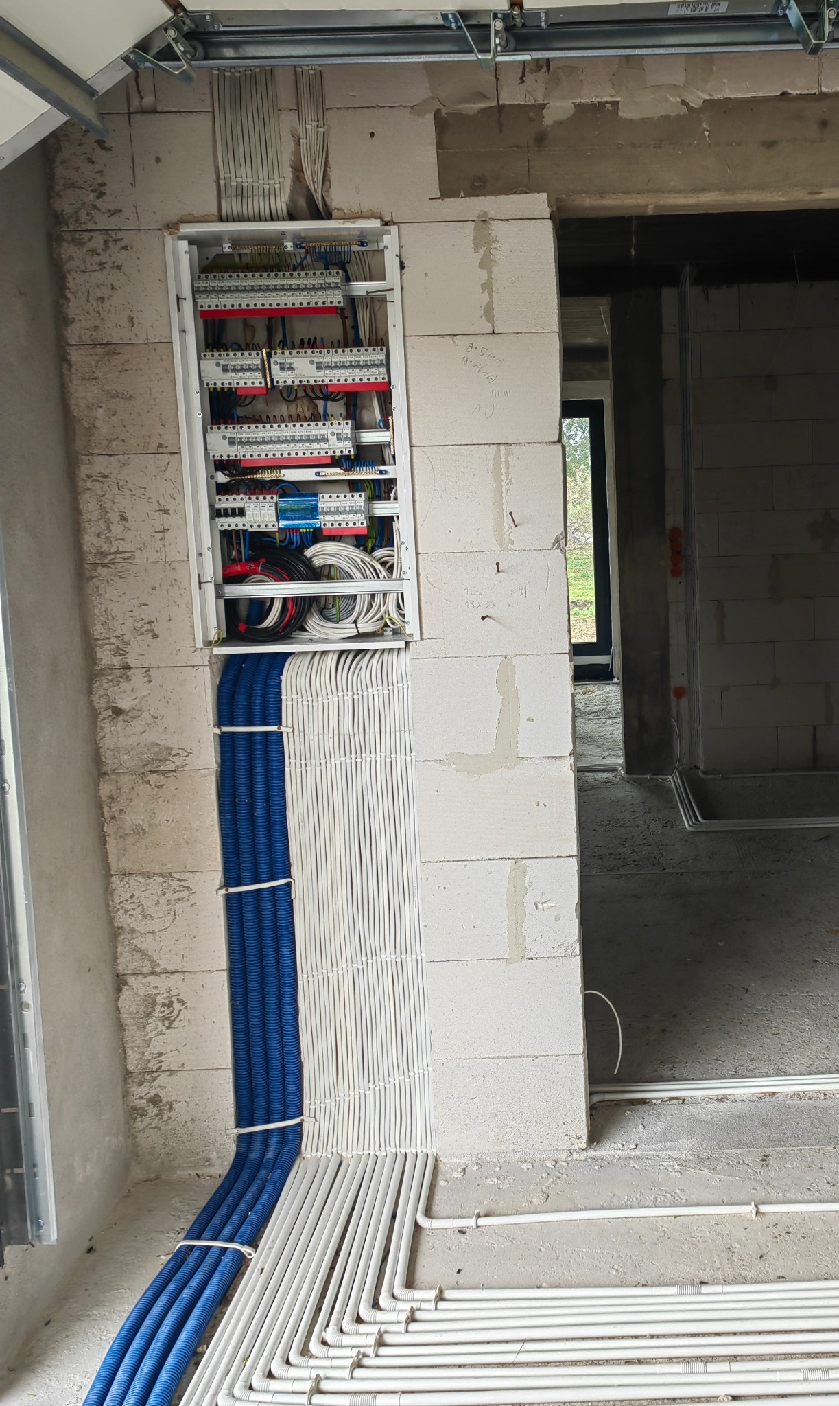 Rozdzielnia elektryczna z licznymi przewodami w białych i niebieskich peszlach, zamontowana w surowej ścianie z betonu komórkowego w trakcie budowy.