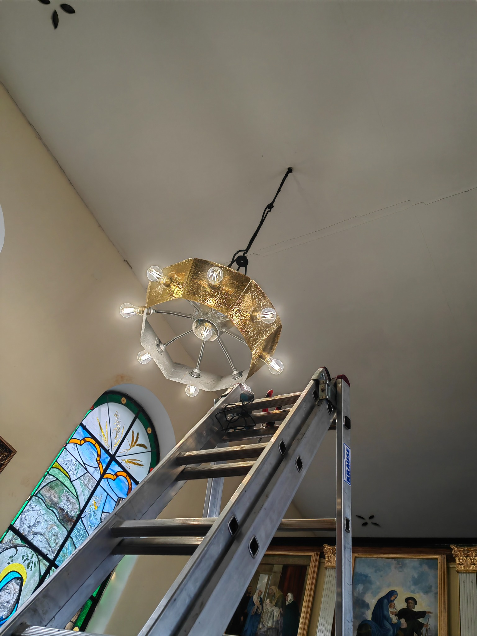 Metalowa drabina oparta o ścianę kościoła w Wardęgowie, z której zwisa złoty, wieloramienny żyrandol z żarówkami Edisona. W tle witraż i obrazy.