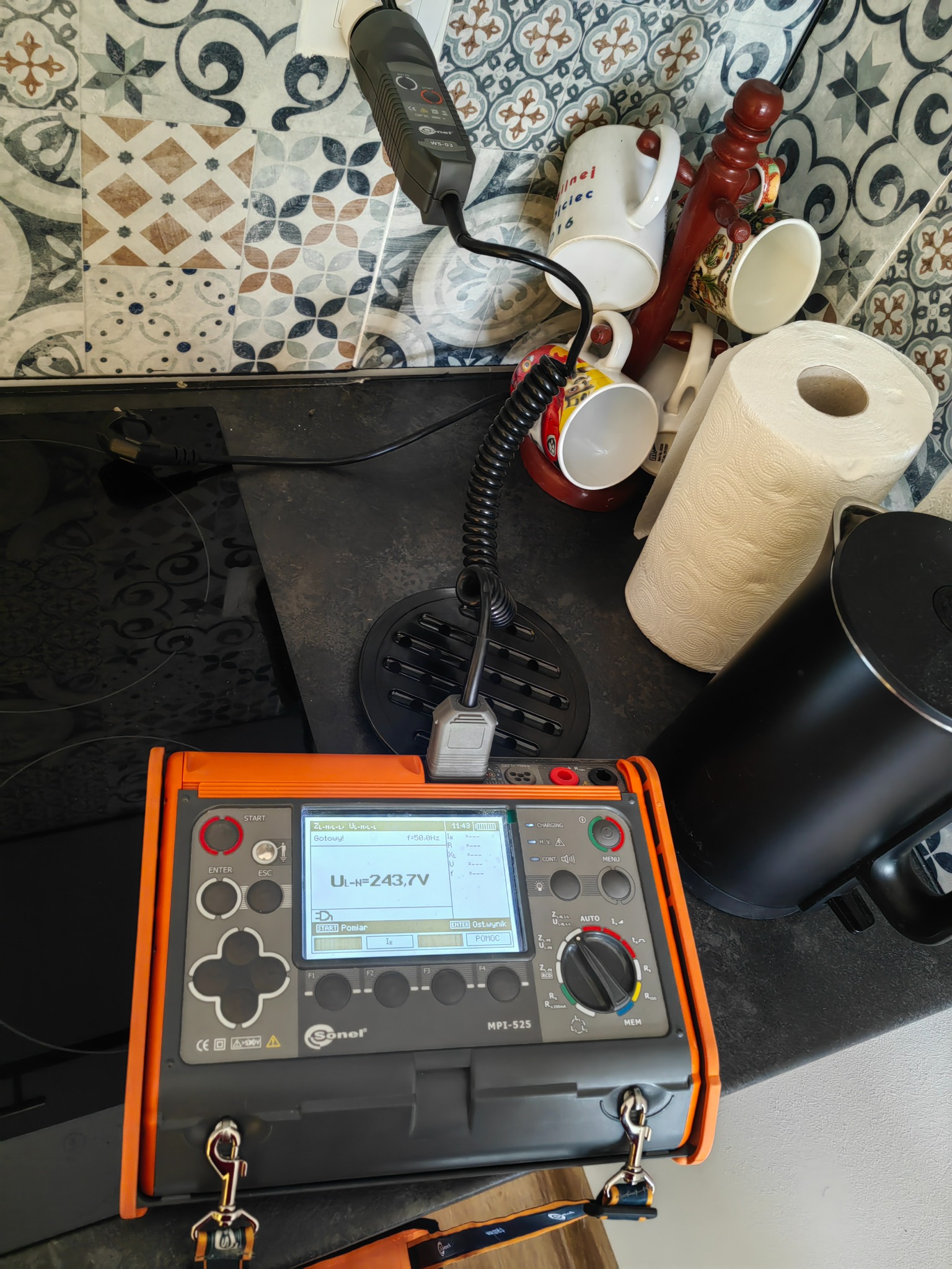 Pomarańczowo-szary miernik elektryczny Sonel MPI-525 z wyświetlaczem wskazującym napięcie 243.7V, ustawiony na czarnym blacie kuchennym obok czajnika i pojemnika na ręczniki papierowe, w tle...