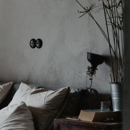 Jola Igielska - Minimalistyczna sypialnia: łóżko z jasną pościelą, drewniana szafka nocna z książką i lampką, roślina w metalowej doniczce na tle ściany z włącznikami światła.