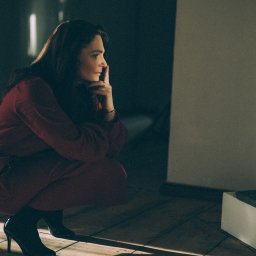 Jola Igielska - Kobieta w czerwonym kombinezonie kuca na drewnianej podłodze, opierając brodę na dłoni, zamyślona, w czarnych szpilkach.