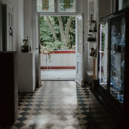 Jola Igielska - Wnętrze domu z otwartymi drzwiami na balkon, podłoga w szachownicę, witraże w oknach.