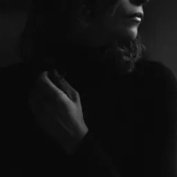 Czarno-białe portretowe ujęcie kobiety z delikatnym makijażem, dłoń dotyka szyi, skierowana w prawo, w ciemnym otoczeniu.