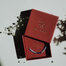 Jola Igielska - Bransoletka z niebieskich i opalizujących koralików w otwartym, czerwonym pudełku marki Ania Kruk, delikatne białe kwiaty i cienie na białym tle.