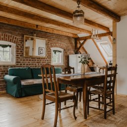 Jola Igielska - Rustykalne wnętrze: ceglana ściana, drewniany strop, zielona sofa, stół z bukietem bzu i okrągły jutowy dywan.
